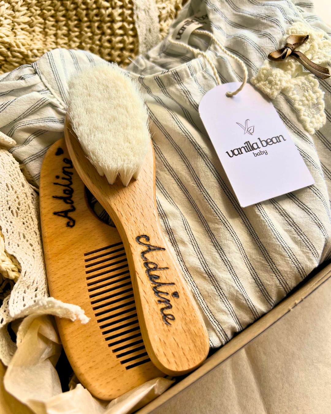 A cosy gift box featuring a Vanilla Bean Baby striped romper, a personalized wooden baby hairbrush and comb set engraved with the name "Adaline", and soft crochet accessories. The Vanilla Bean Baby tag is attached to the romper, emphasising the brand's minimalist and sentimental aesthetic.